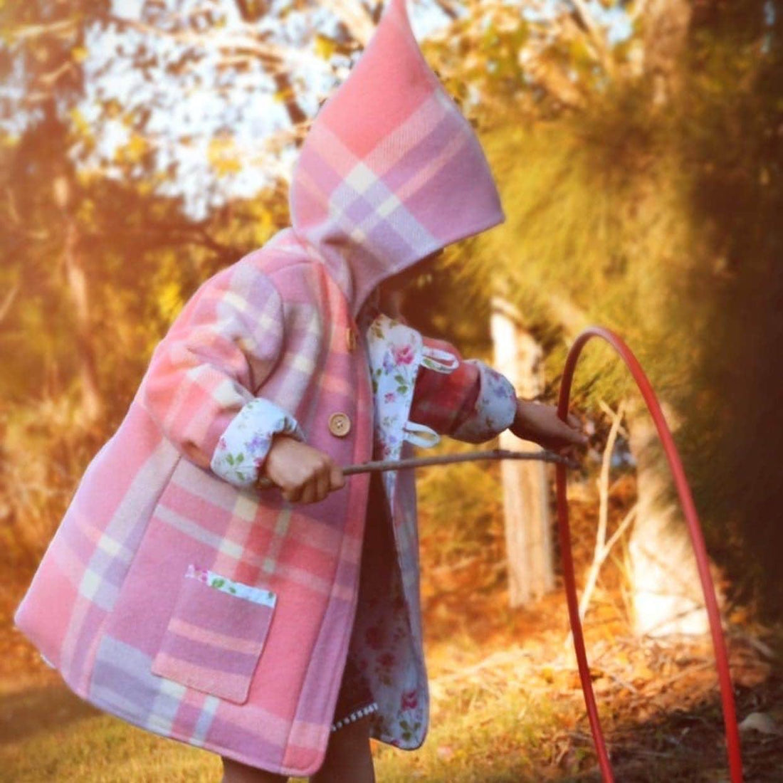 Photo features a little girl wearing a pink woollen jacket with a pixie hood playing outside in a garden