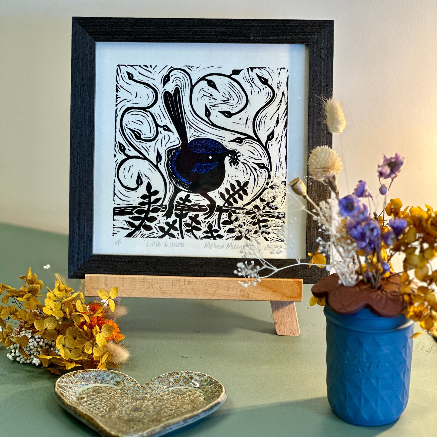 Blue wren art work called little lunch by Melissa Murry framed and sitting on a sideboard with a vase of flowers.