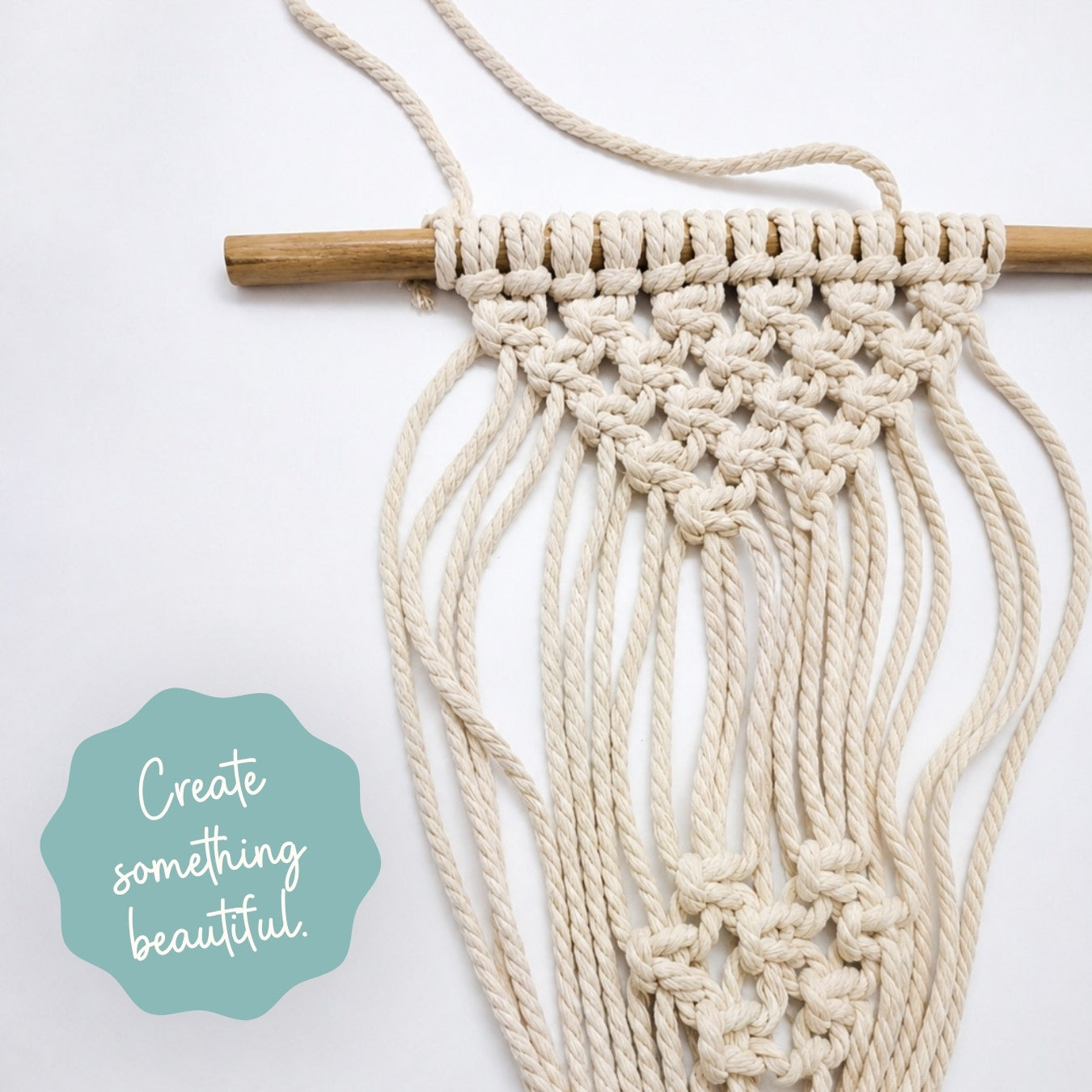 A cream macrame wall hanging with a wooden dowel, shown against a white background with a teal badge that reads 'Create something beautiful.'.